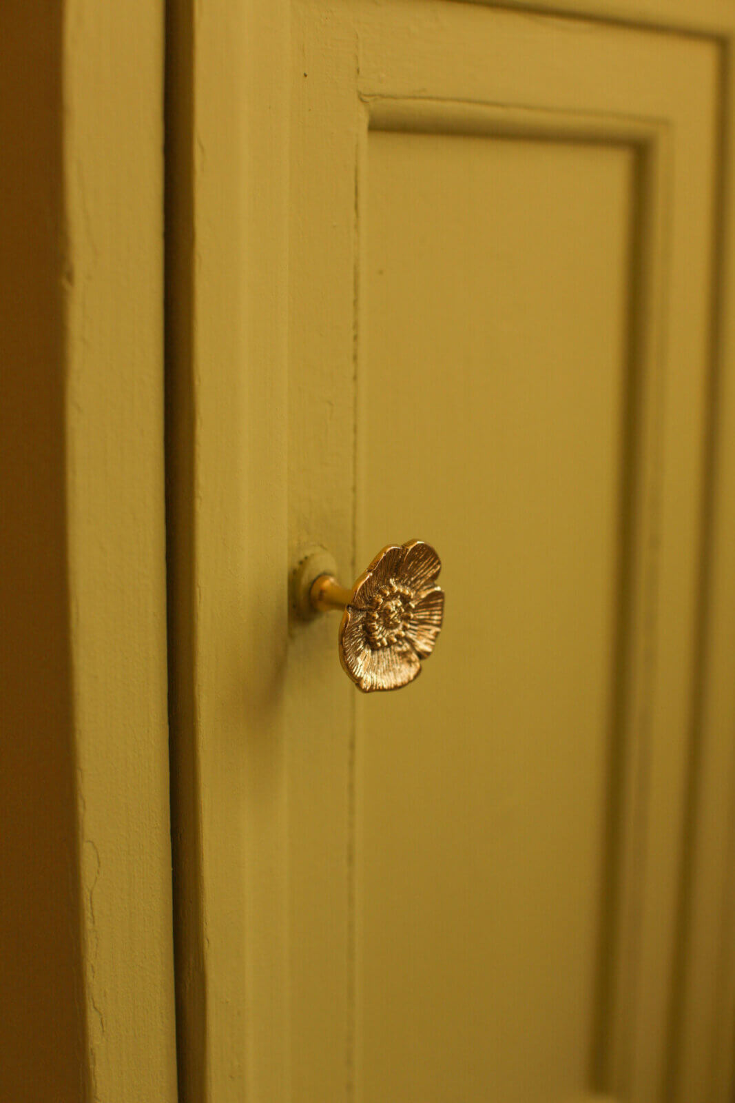 A close-up of a beige cabinet door featuring the Mia Poppy Knob, shaped like a gold ginkgo leaf. The cabinet has a subtly textured finish and recessed panel detailing.